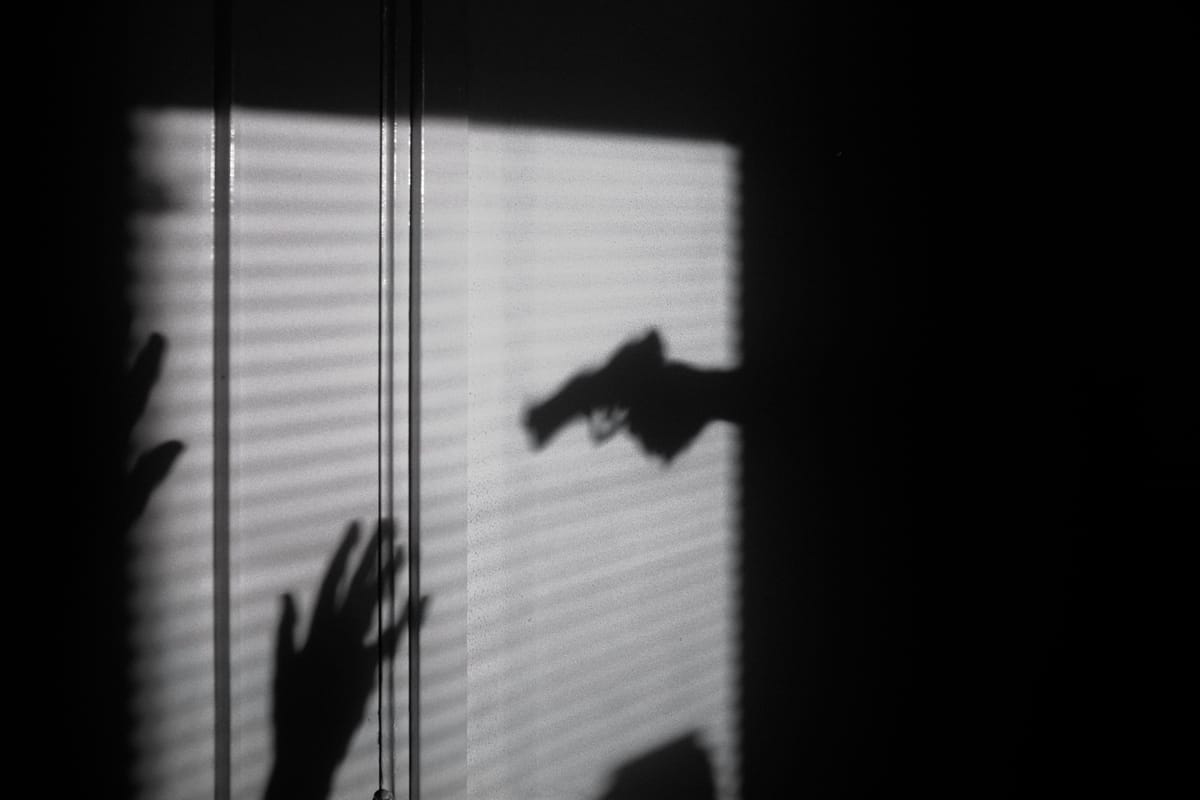 On a wall, the shadow of a hand holding a gun meets the shadow of a hand raised in defense.
