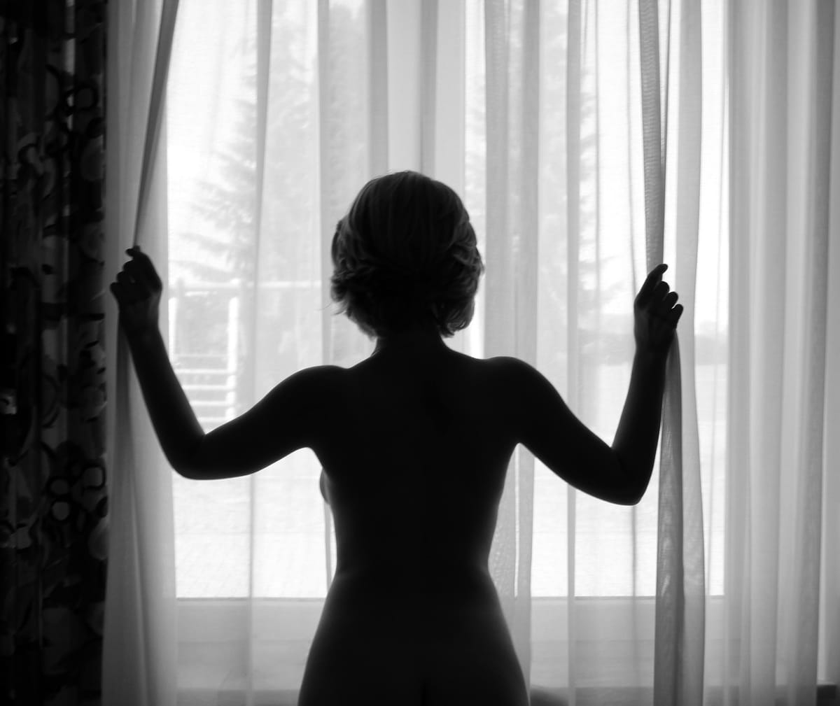一位女性的黑白剪影，雙手拉開白色窗簾，逆光站立