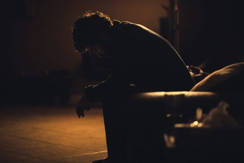 A person hunched in darkness, head bowed under warm dim light, surrounded by shadows