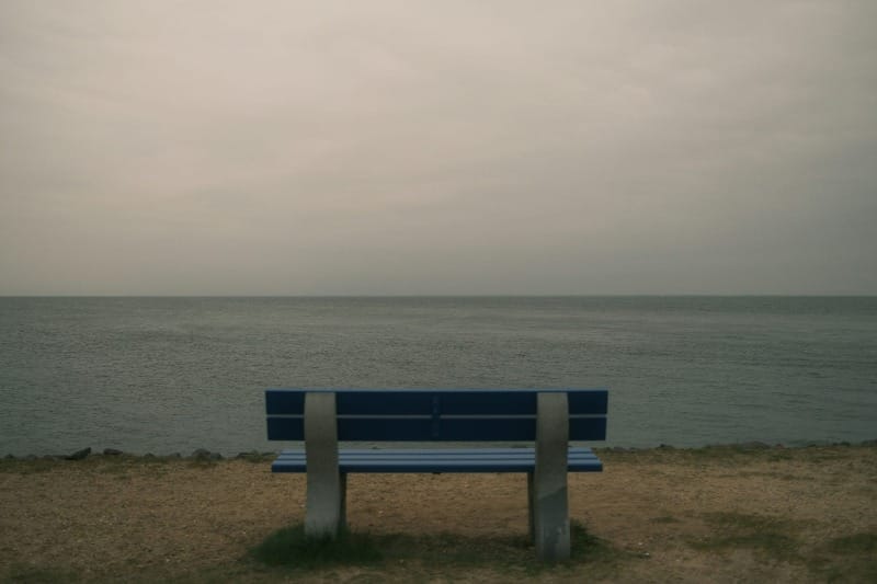 A lone blue bench facing a calm grey sea under an overcast sky
