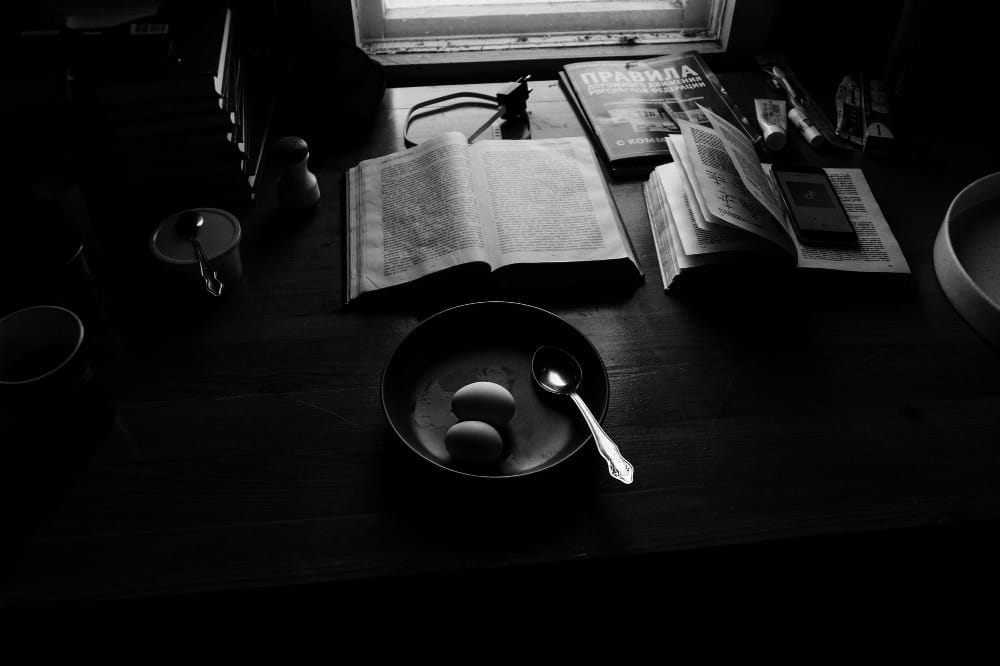 Black and white photo of a messy desk with open books and a bowl of eggs, symbolizing the paralysis of procrastination and the moment before starting.